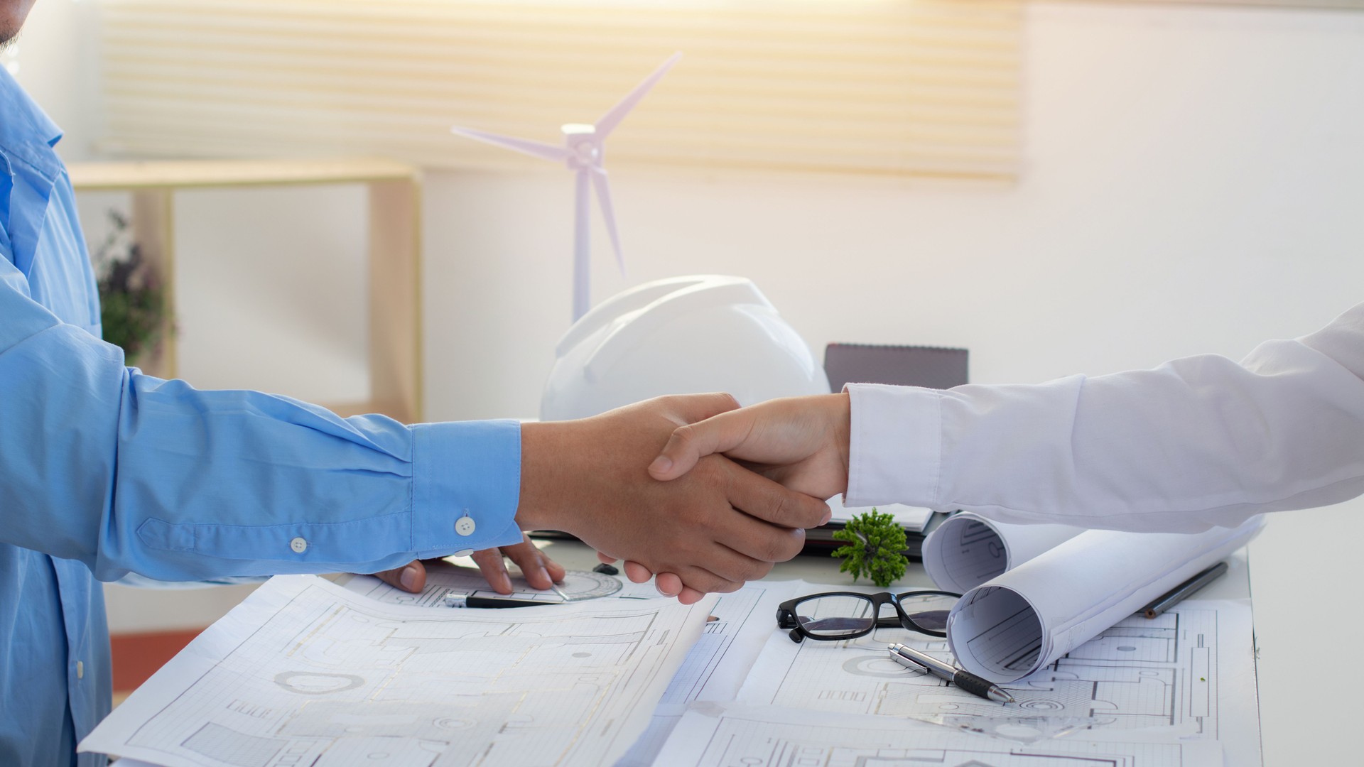 Two people in dress shirts shake hands over architectural blueprints, signifying an agreement or partnership. A hardhat and miniature wind turbine suggest a construction or renewable energy project.